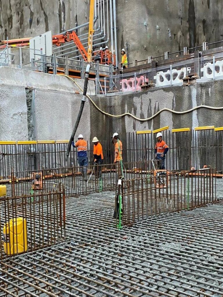 Construction workers in high-visibility orange gear and white helmets are working on a site with steel rebar grids and concrete walls. They appear to be preparing for a concrete pour, with one worker using a hose-like tool. Scaffolding and safety barriers are visible.