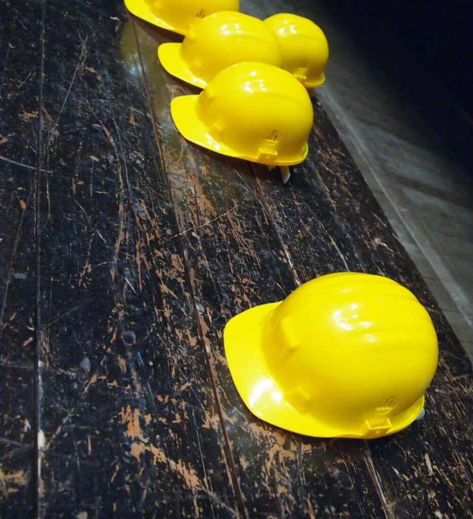 Four yellow construction helmets placed in a line on a worn, dark wooden surface. The helmets reflect light and create a contrast against the rough texture of the wood. The scene appears to be on a construction site or industrial setting.
