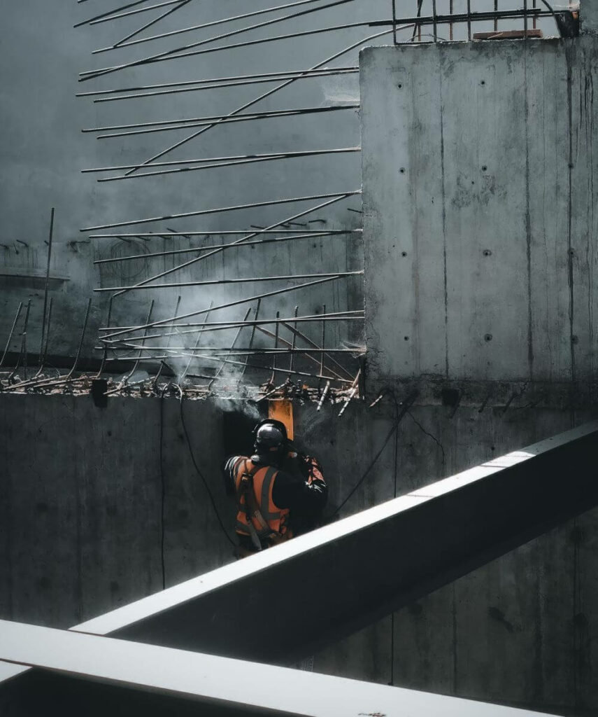 A construction worker in an orange safety vest and helmet welds metal beams on a partially built concrete structure. Sparks fly from the welding torch, illuminating the surrounding area in the dim light. Reinforced steel rods jut out from the walls and beams.