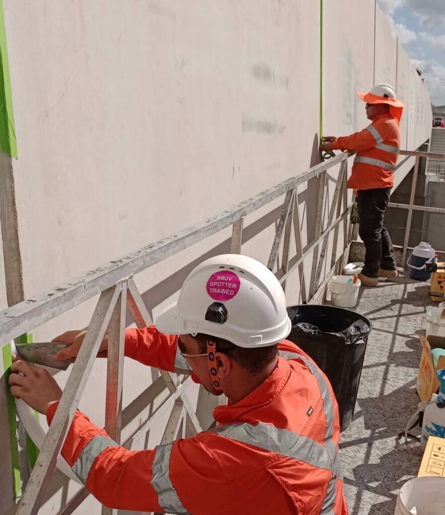 Two construction workers wearing orange high-visibility clothing and white hard hats are working on a structure. One worker in the foreground is standing close to a metal framework, while the other is standing further back near a concrete wall.