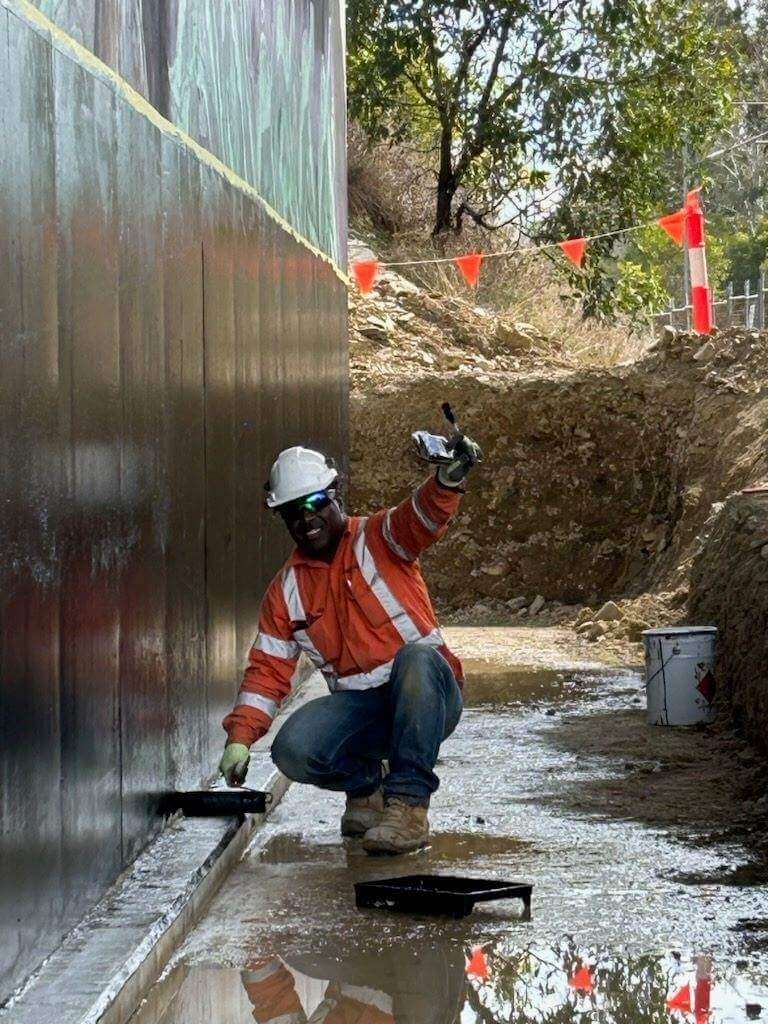 A construction worker in an orange and white safety jacket and white helmet kneels next to a large wall, applying material with a trowel. He smiles and gives a thumbs-up with his other hand. The ground is muddy, and there are safety flags and trees in the background.