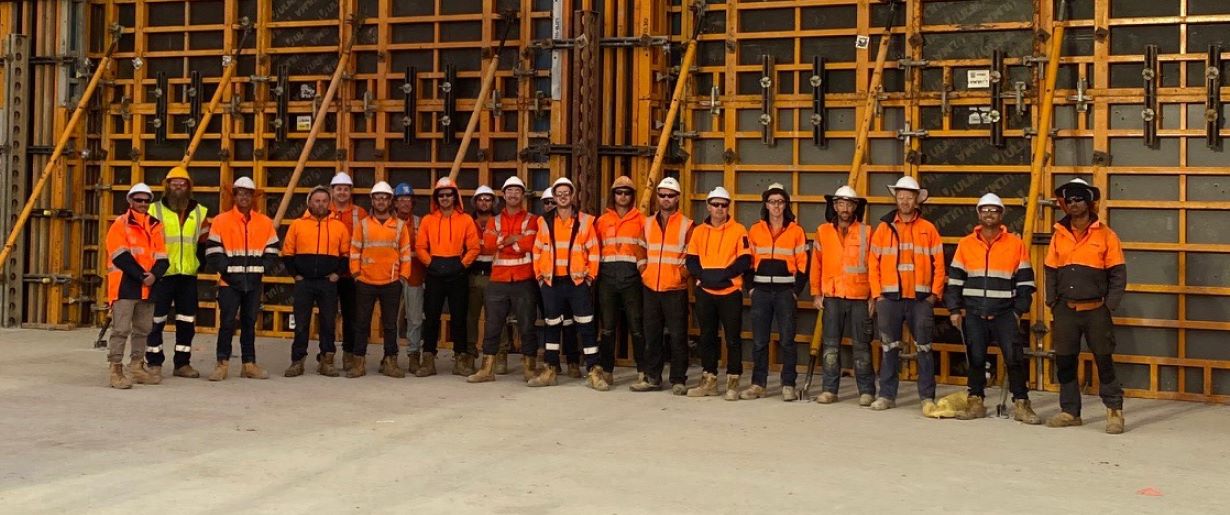 A group of 18 construction workers from About Ground One Civil, dressed in bright orange and yellow safety vests, hard hats, and work boots, stand proudly in front of a large construction site. They appear to be posing for a group photo on the concrete floor.