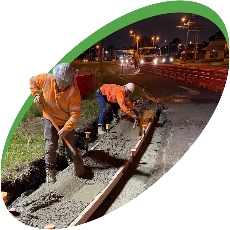 Workers in orange safety vests and hard hats spread concrete with shovels at a night construction site. Orange traffic barriers line the road with vehicles seen at a distance. The scene is framed by a green, curved line.