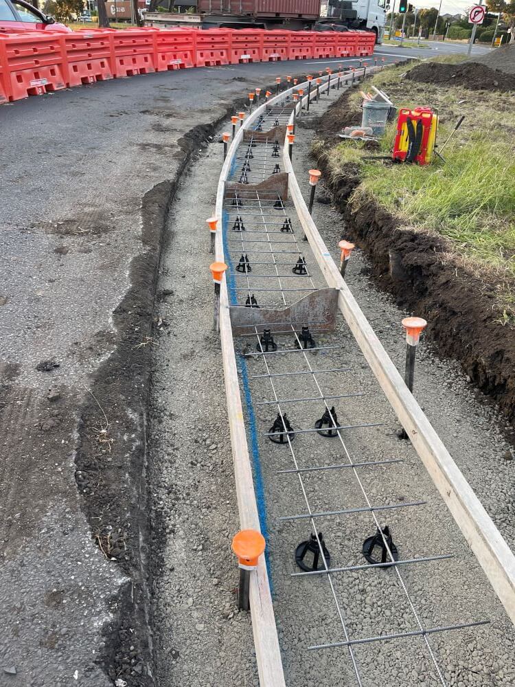 A construction site with groundwork for a curving sidewalk or road. Rebar is laid out within wooden frames along the curve, with plastic caps on exposed steel rods. Red barriers line the site, and green grass is visible on the right. Construction tools are nearby.