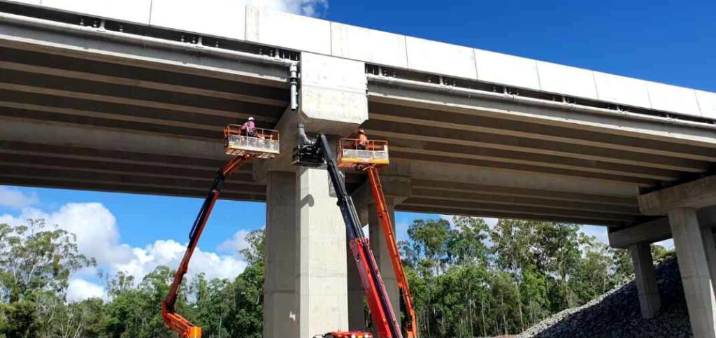 Two workers are on elevated platforms, inspecting or repairing the underside of a large concrete bridge. The platforms are supported by long, mechanical arms. Below the bridge, there's a forested area with trees and a pile of rocks. The sky is clear and blue.
