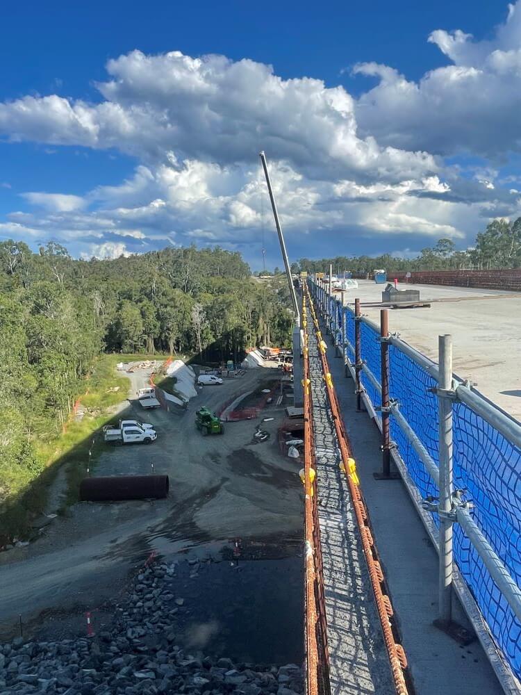 A construction site with a long bridge under development. The bridge is lined with blue safety netting on one side. Construction vehicles, equipment, and materials are scattered below on a dirt road. The background features green trees and a partly cloudy sky.