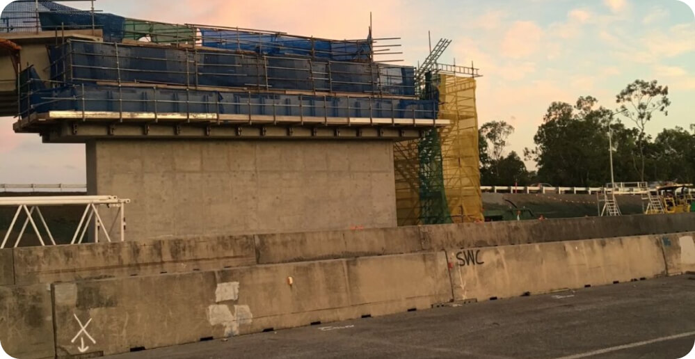 An elevated section of a bridge under construction with scaffolding and blue tarps showcases ongoing services. Concrete barriers separate the construction area from the road. Trees are visible in the background, and the sky has a pinkish hue, suggesting either sunrise or sunset.