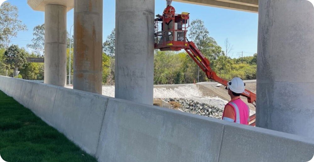Two construction workers wearing hard hats and safety vests provide essential services under a bridge. One operates a cherry picker lift, while the other stands on the ground observing. The scene is set outdoors with trees and a clear sky in the background.