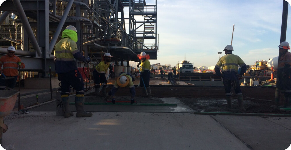 Construction workers in safety gear are diligently providing their services on a concrete slab at an industrial site. The workers are spreading freshly poured concrete, using tools and wearing high-visibility clothing, hard hats, and boots. Machinery and scaffolding are visible in the background.
