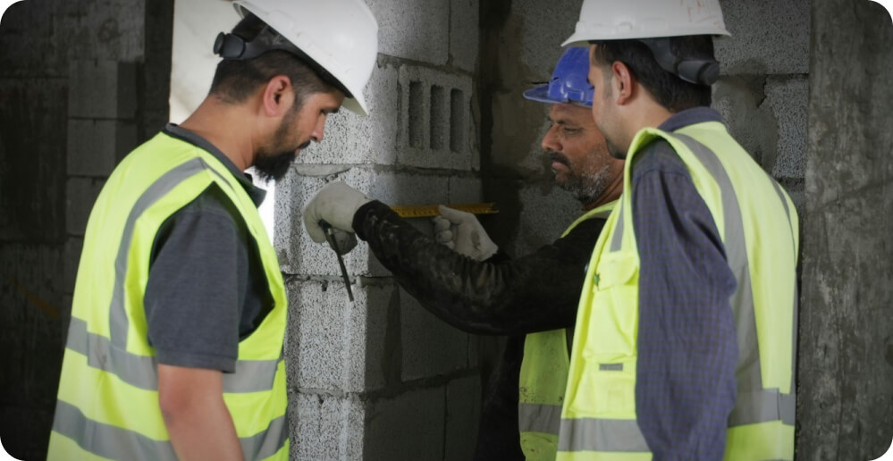 Three construction workers wearing safety helmets and reflective vests are measuring a wall made of concrete blocks. One worker holds a measuring tape, while the others observe. They are providing essential services in the middle of a building under construction.