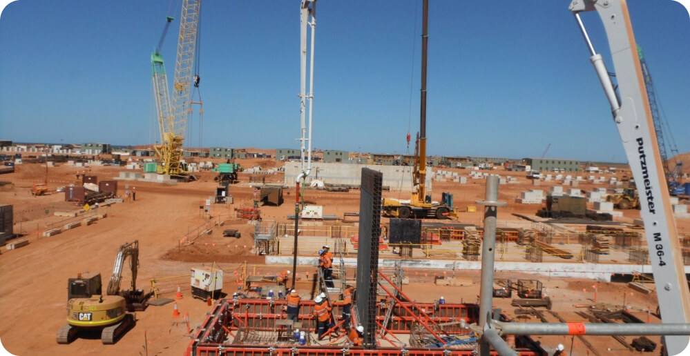 Construction site with cranes, machinery, and workers assembling steel structures on a sandy landscape under a clear blue sky. Residential buildings, benefiting from our construction services, are visible in the background.