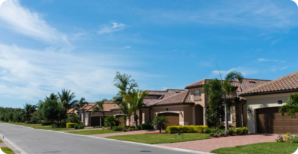 A suburban street with several single-family homes featuring Spanish-style architecture, including terracotta roofs and stucco exteriors. Well-maintained lawns and palm trees enhance the serene neighborhood under a clear blue sky. The street is empty of vehicles but lined with necessary services for residents.