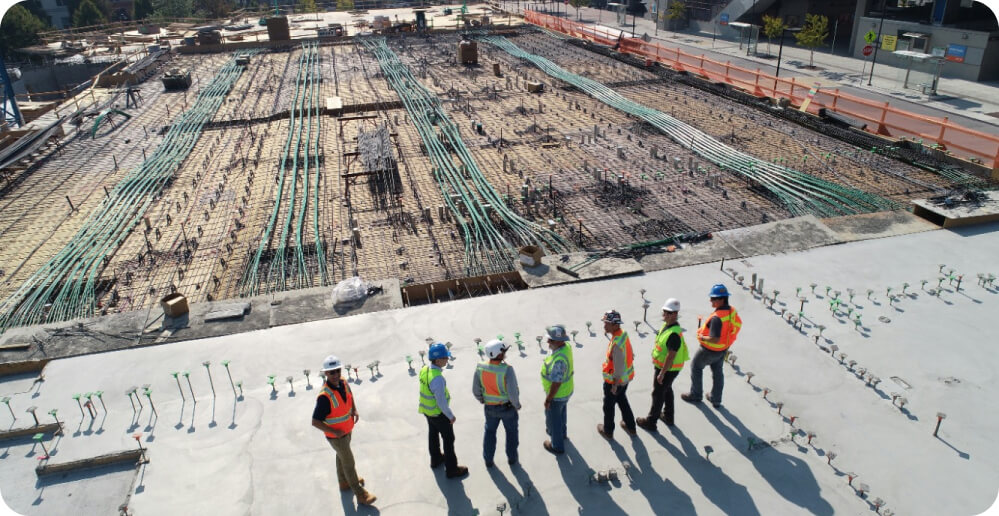 Six construction workers in safety gear and helmets stand on a concrete area, discussing the ongoing services on a large construction site. The site is filled with an array of rebar and conduits laid out across the expanse, showcasing the early stages of building infrastructure.