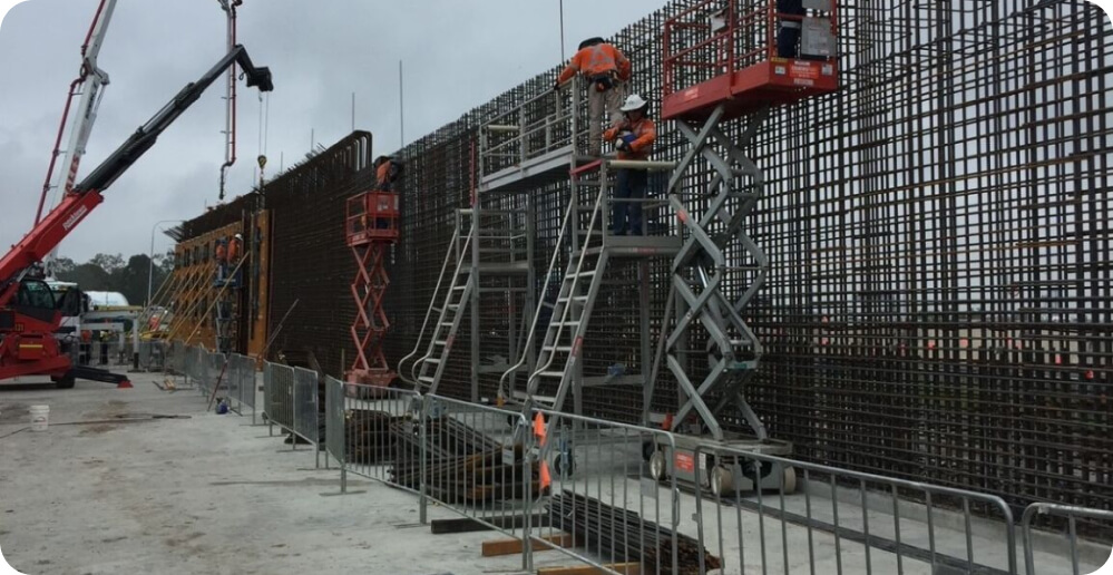 Construction workers in safety gear diligently provide services on a large metal rebar structure. Some workers are on elevated platforms, while others operate from the ground. The construction site is surrounded by barriers, with heavy machinery in use under an overcast sky.