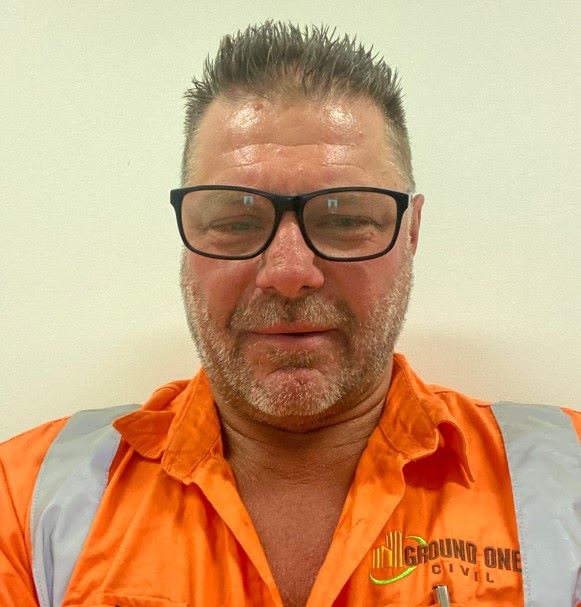 A man with short hair and glasses is wearing an orange shirt with reflective stripes and a logo that reads "Ground One Civil." He is standing in front of a plain light-colored background, looking at the camera with a slight smile.