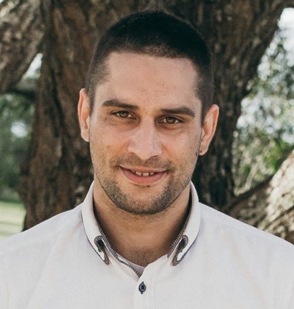 A man with short dark hair and a trimmed beard is standing outdoors in front of a large tree. He is wearing a white button-up shirt and is smiling at the camera. The background is slightly blurred, featuring green foliage.