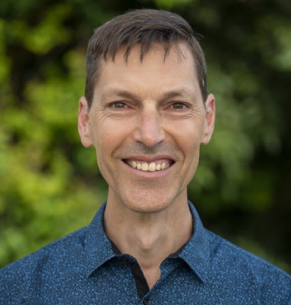 A man with short brown hair and wearing a blue button-up shirt smiling at the camera. The background is blurred greenery.