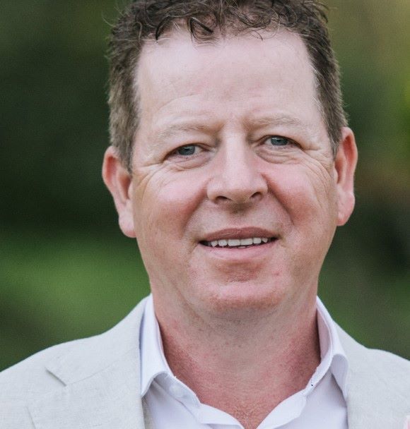 A man with short curly brown hair and blue eyes is smiling at the camera. He is wearing a light-colored blazer over a white button-up shirt. The background is a blur of natural greenery.